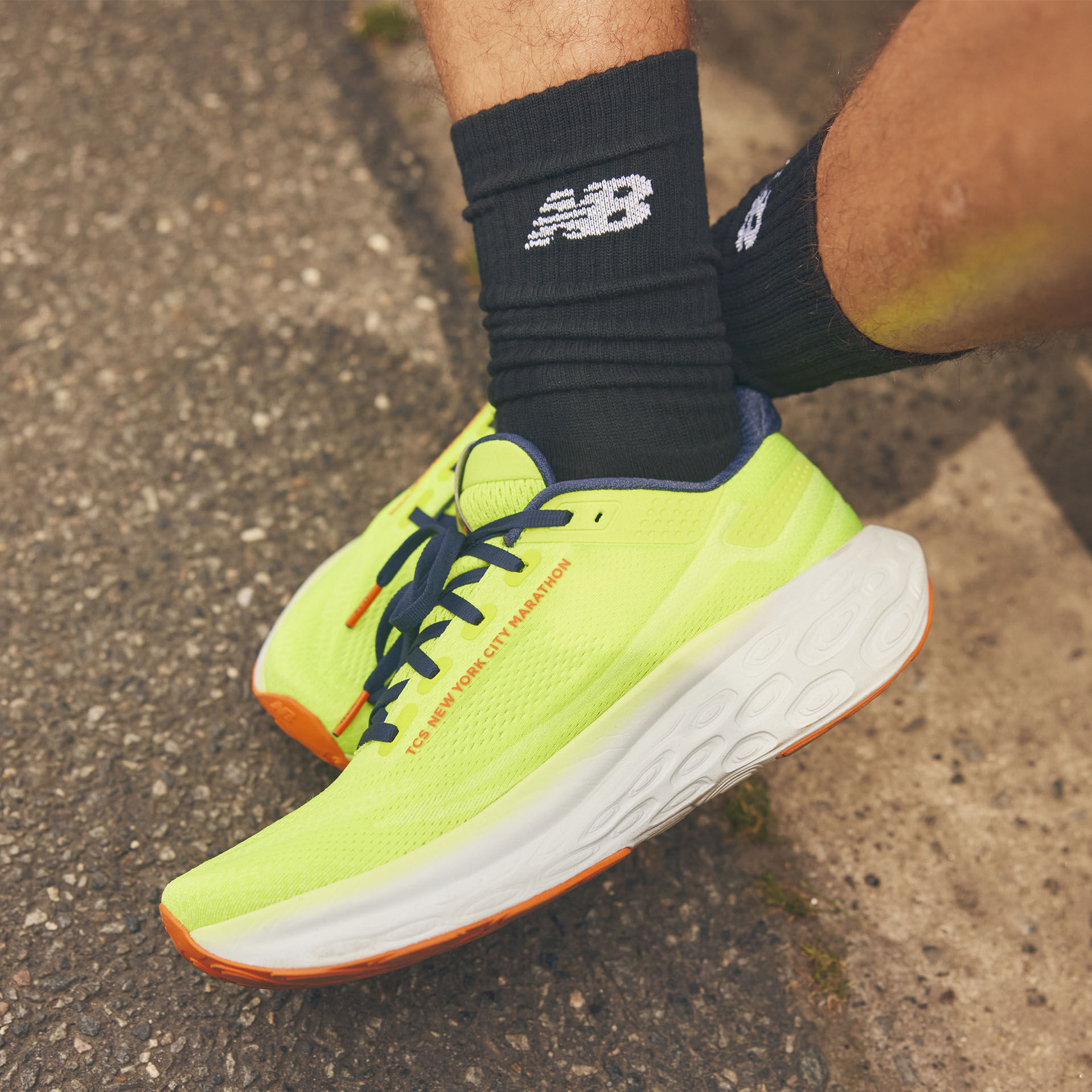Imagen de la rodilla hacia abajo de un hombre usando el calzado New Balance Fresh Foam X 1080 en color amarillo brillante y naranja con calcetines deportivos contra un fondo de concreto 