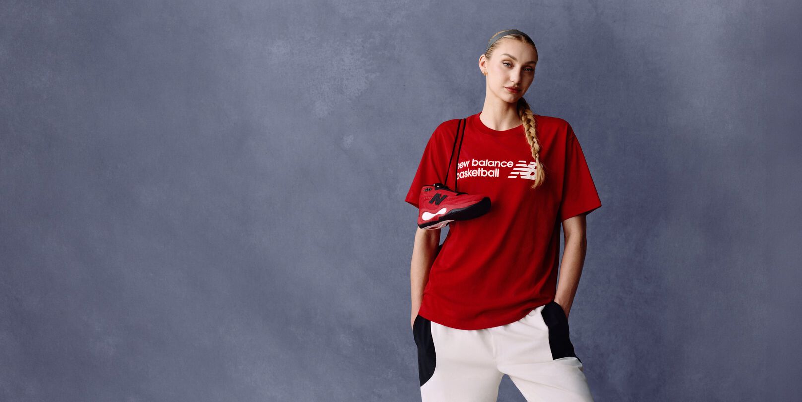 Tenis de baloncesto New Balance en rojo y negro sobre fondo gris claro, posicionado para destacar el perfil lateral con el logo &ldquo;N&rdquo; negro y el detalle blanco de la mediasuela.