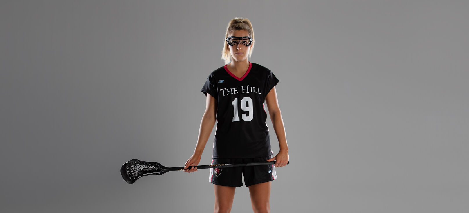 Female lacrosse player dressed in full gear posing in front of a grey studio background 