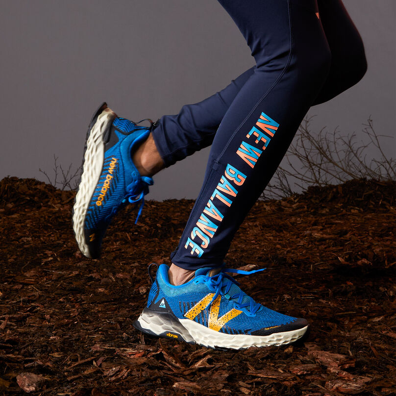 Knee down of person running on trail dirt with wearing navy leggings, showing the side profile of blue Hierro trail running shoe