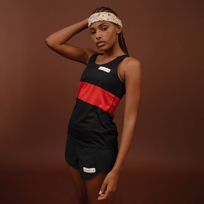 Woman wearing a black tank with a red stripe through the middle, black shorts, and a yellow bandana around her forehead, on a maroon background