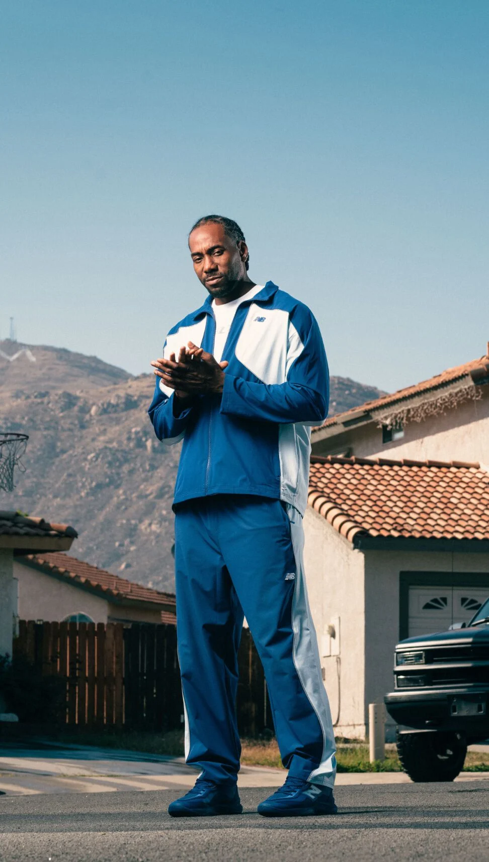 Person wearing a blue and white tracksuit and matching sneakers stands on a suburban street with mountains and houses in the background.