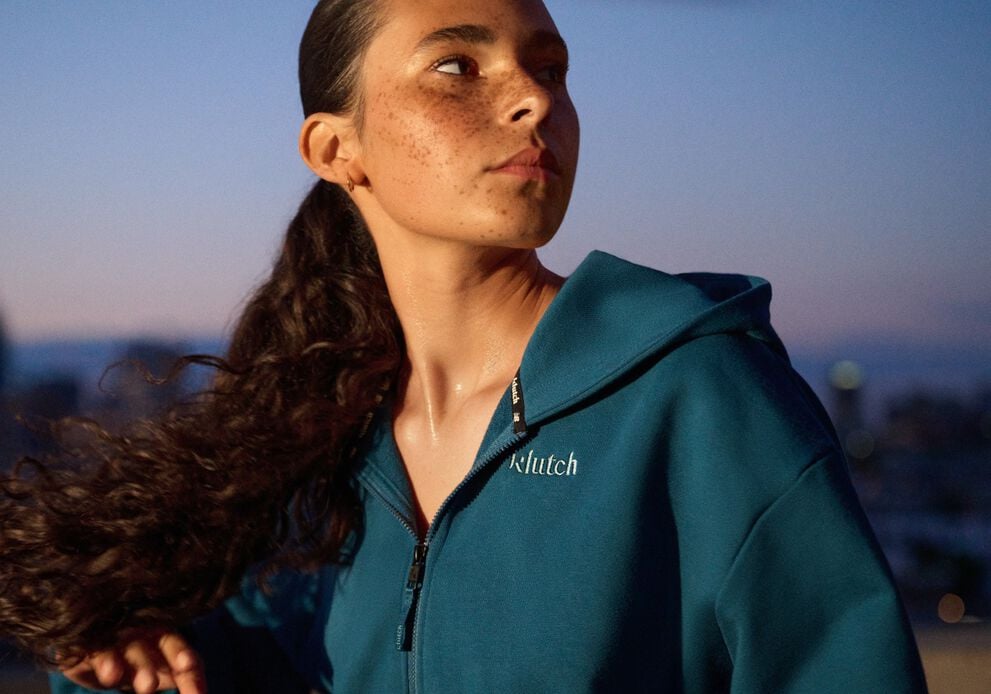 Athlete wearing a teal zip-up hoodie with a city skyline blurred in the background at dusk.
