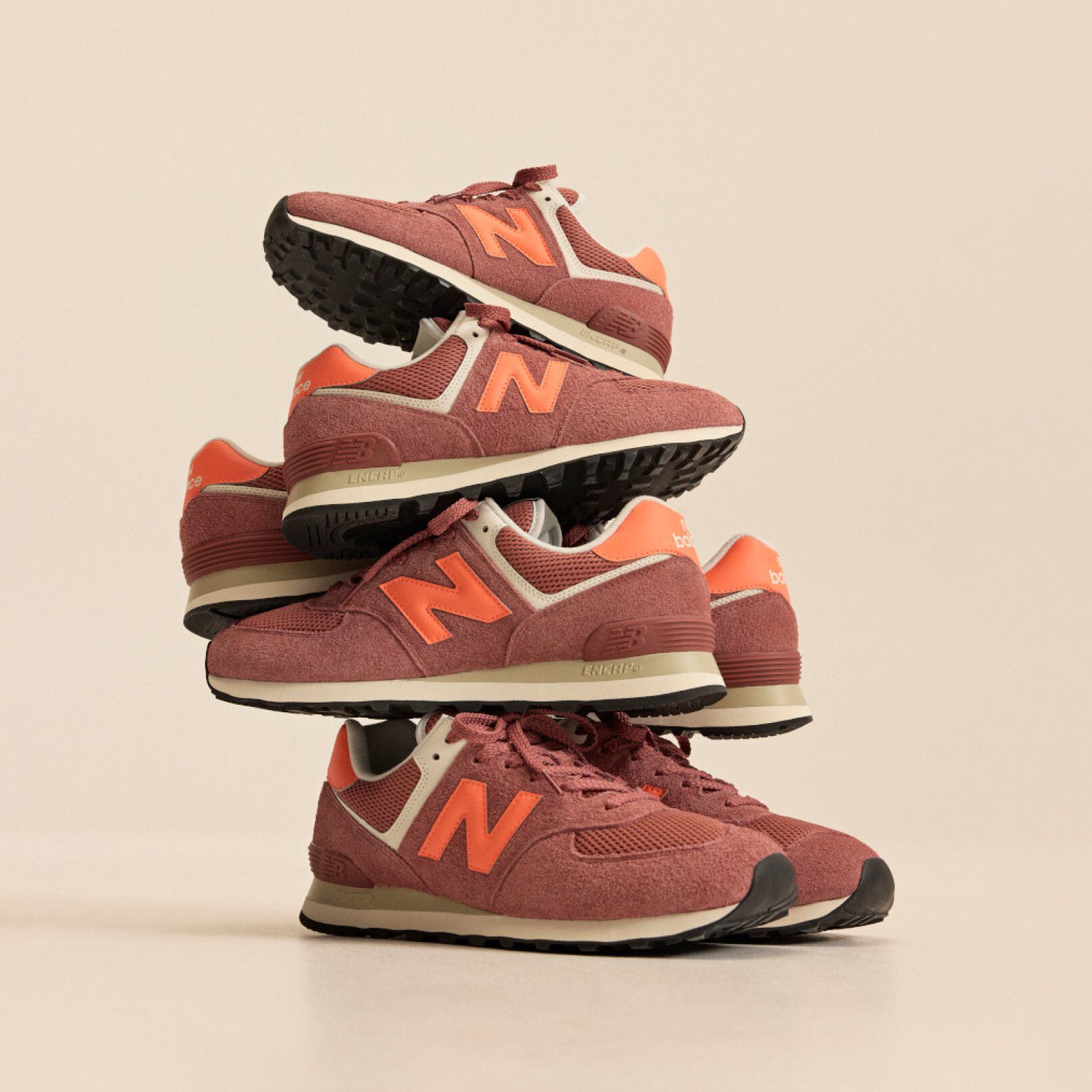 Stack of brick‑red New Balance sneakers with orange accents arranged in a balanced tower against a beige background.