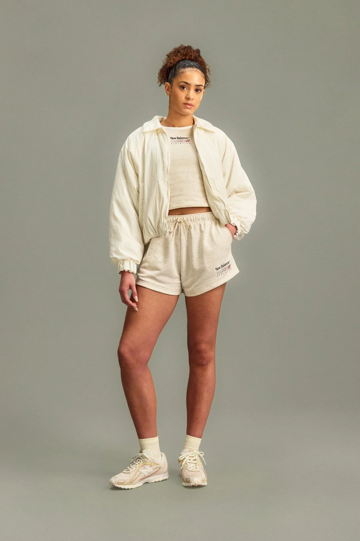 A woman wearing a New Balance shirt, jacket and shorts standing in front of a grey studio background.
