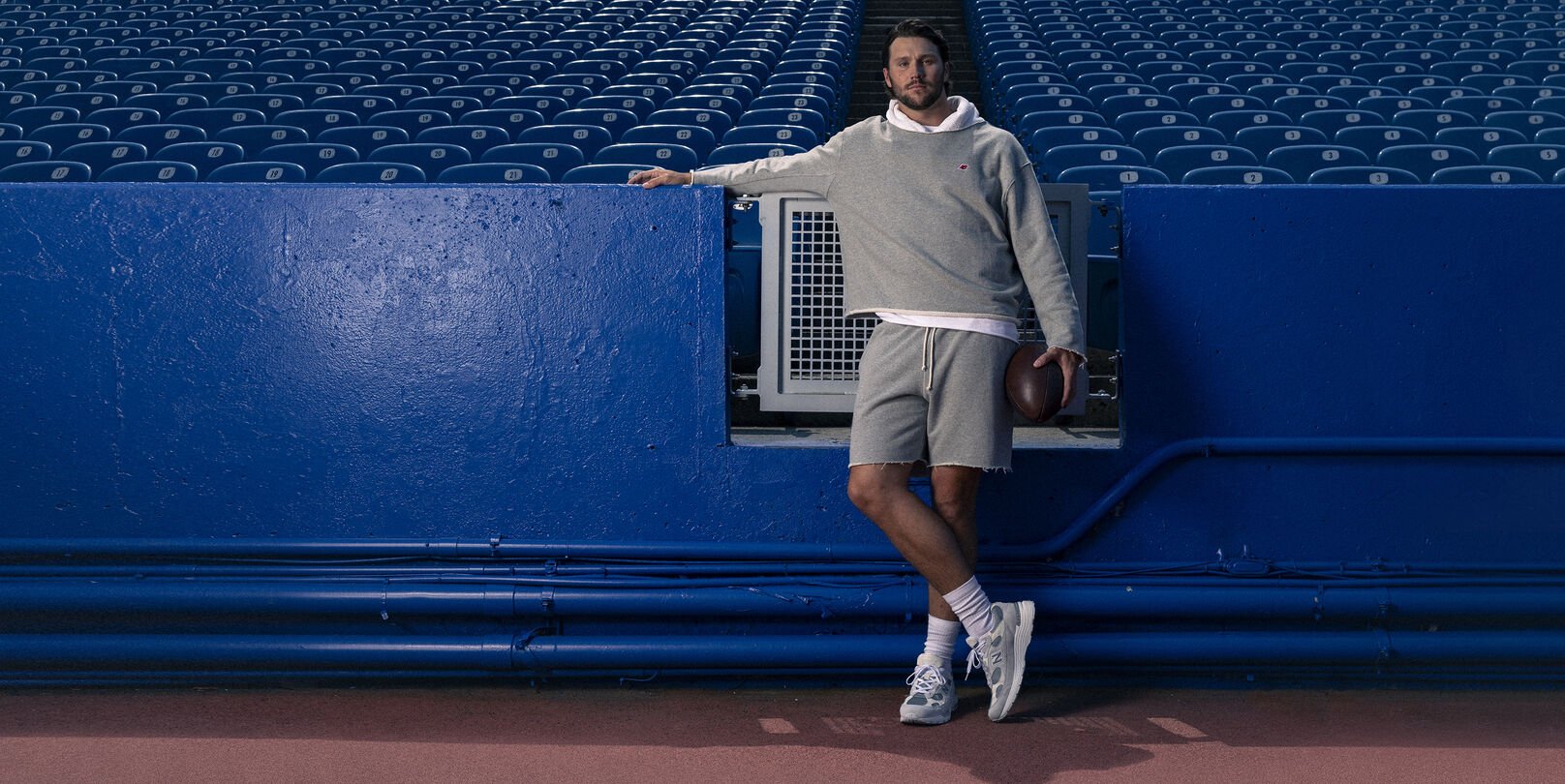 Young male athlete in a grey hoodie leaning on a stadium fence.