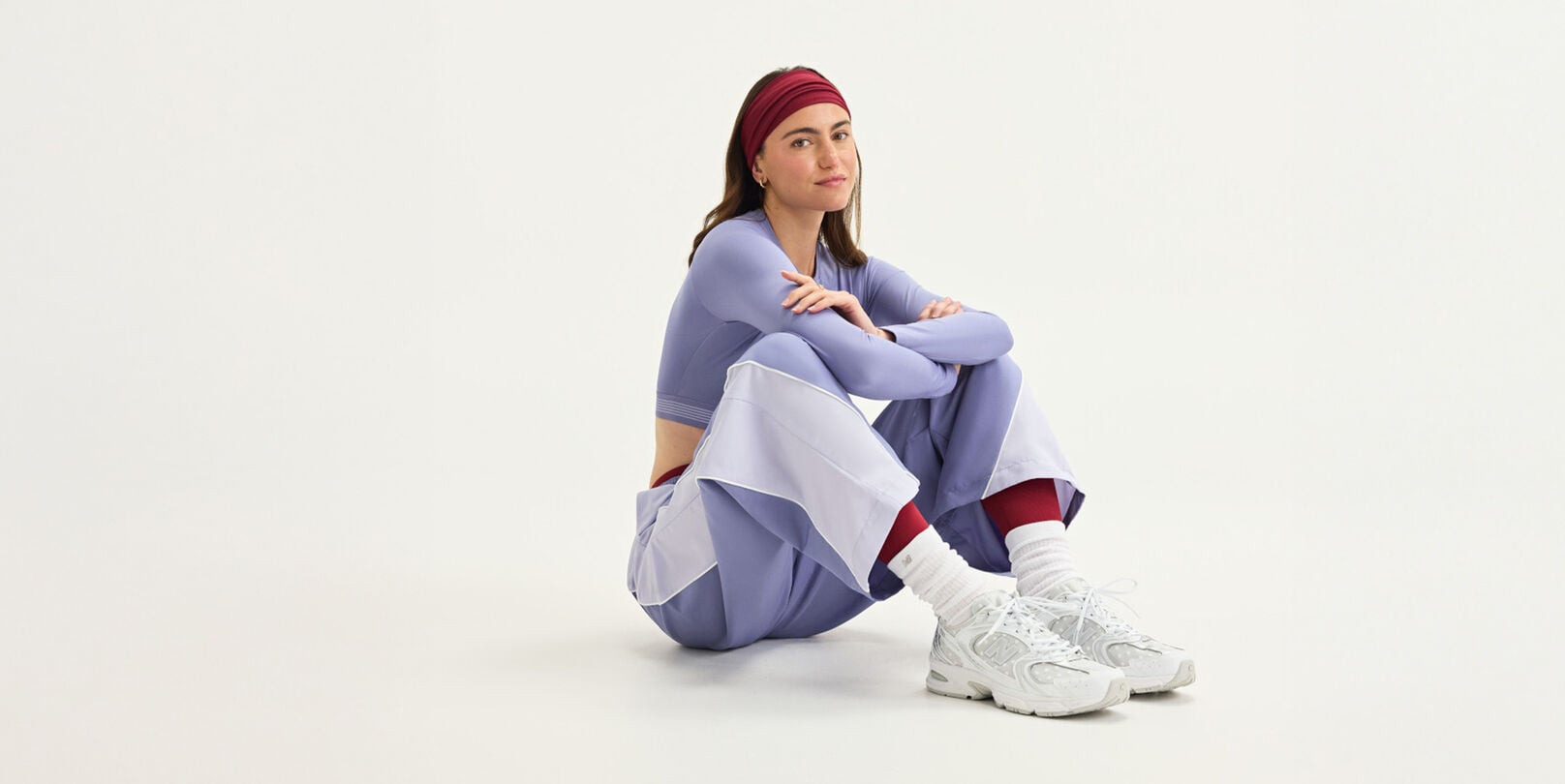 A woman sitting wearing New Balance gear on a white background.