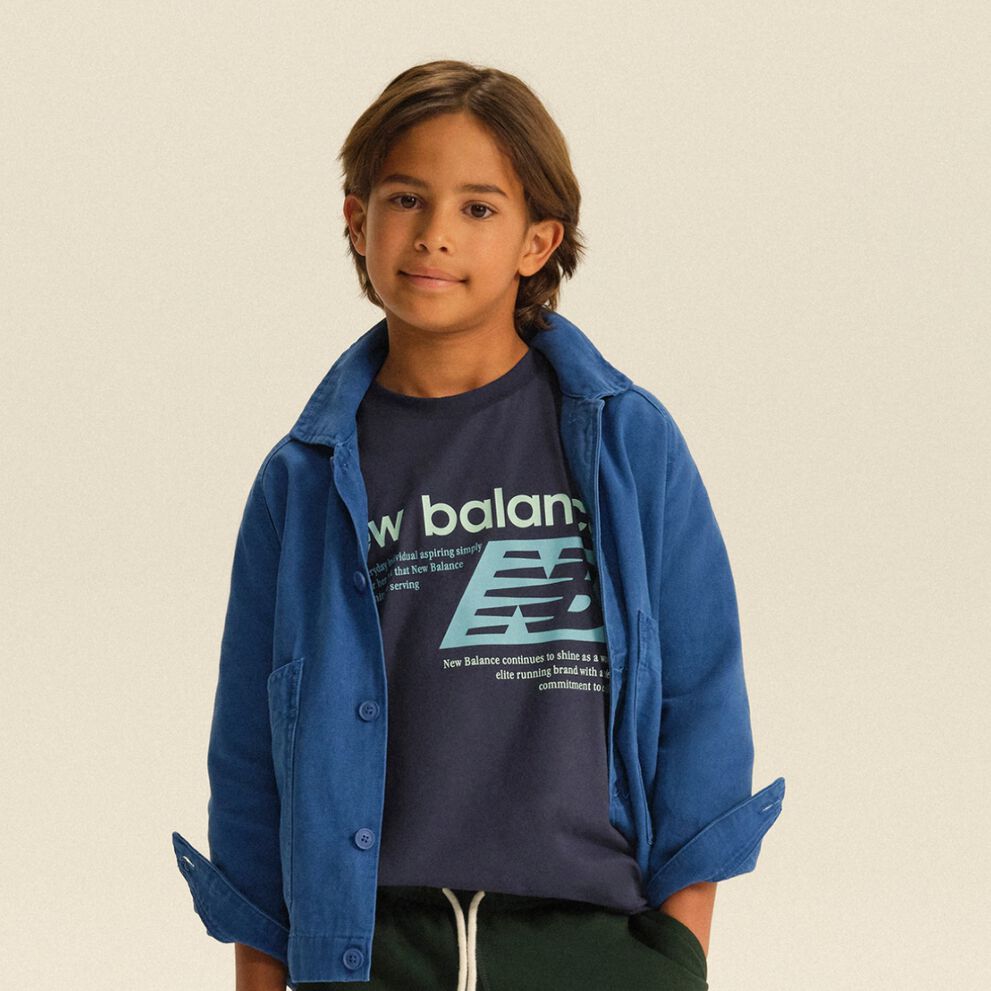 Child wearing a blue overshirt layered over a graphic New Balance T-shirt and dark joggers in a clean studio setting.
