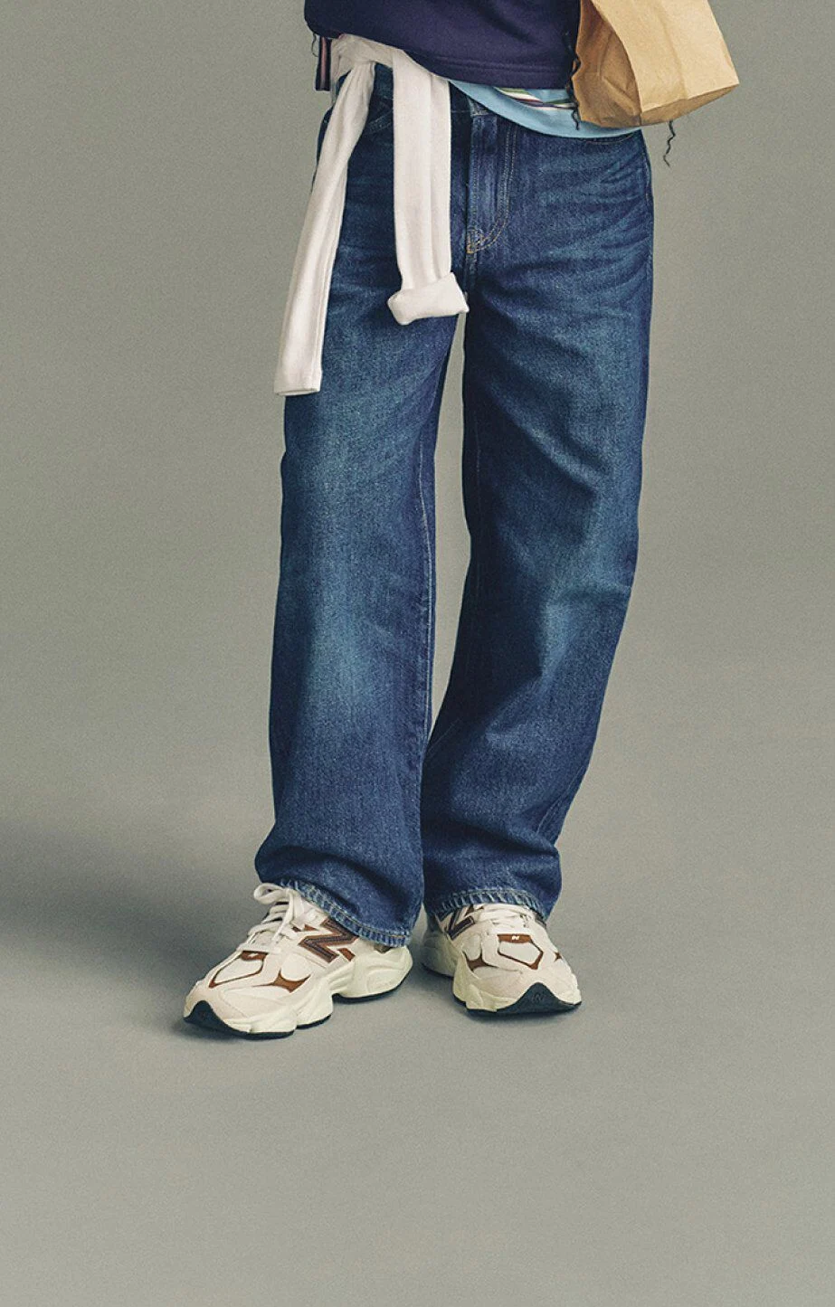 Close-up of a person wearing loose blue jeans and white New Balance sneakers with brown accents, standing against a plain background while holding a paper bag.