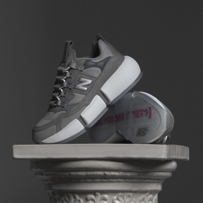 Pair of grey Vision Racer sneakers sitting on a stone pedestal against a dark studio backdrop.