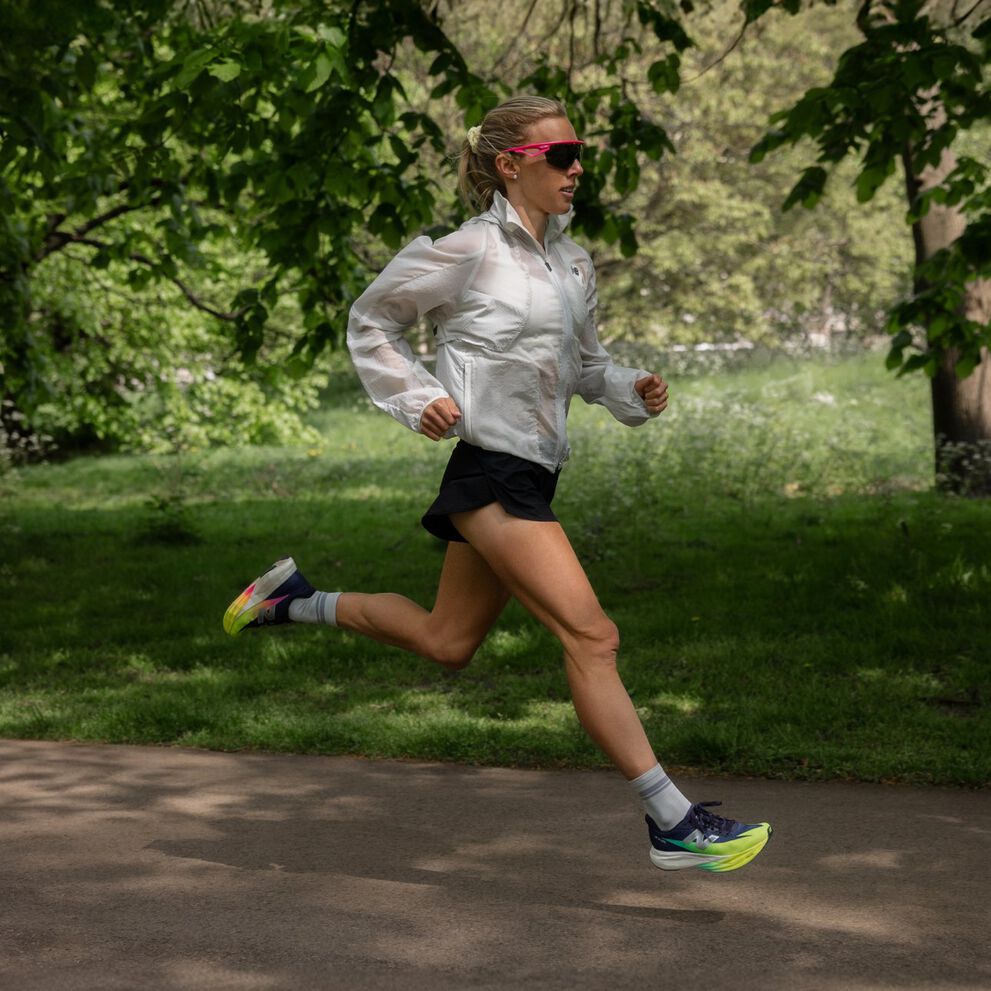 Emily Sisson corriendo al aire libre en un camino pavimentado en un parque usando una chaqueta blanca, pantalones cortos negros y calzado de correr con colores brillantes.&nbsp;