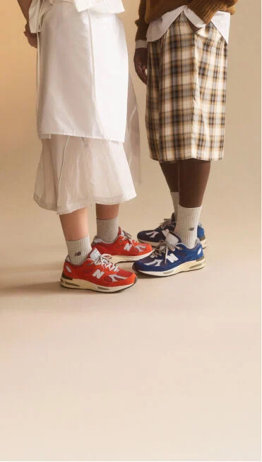 Waist down view of a man and woman wearing New Balance Made in UK sneakers standing in a beige studio environment.