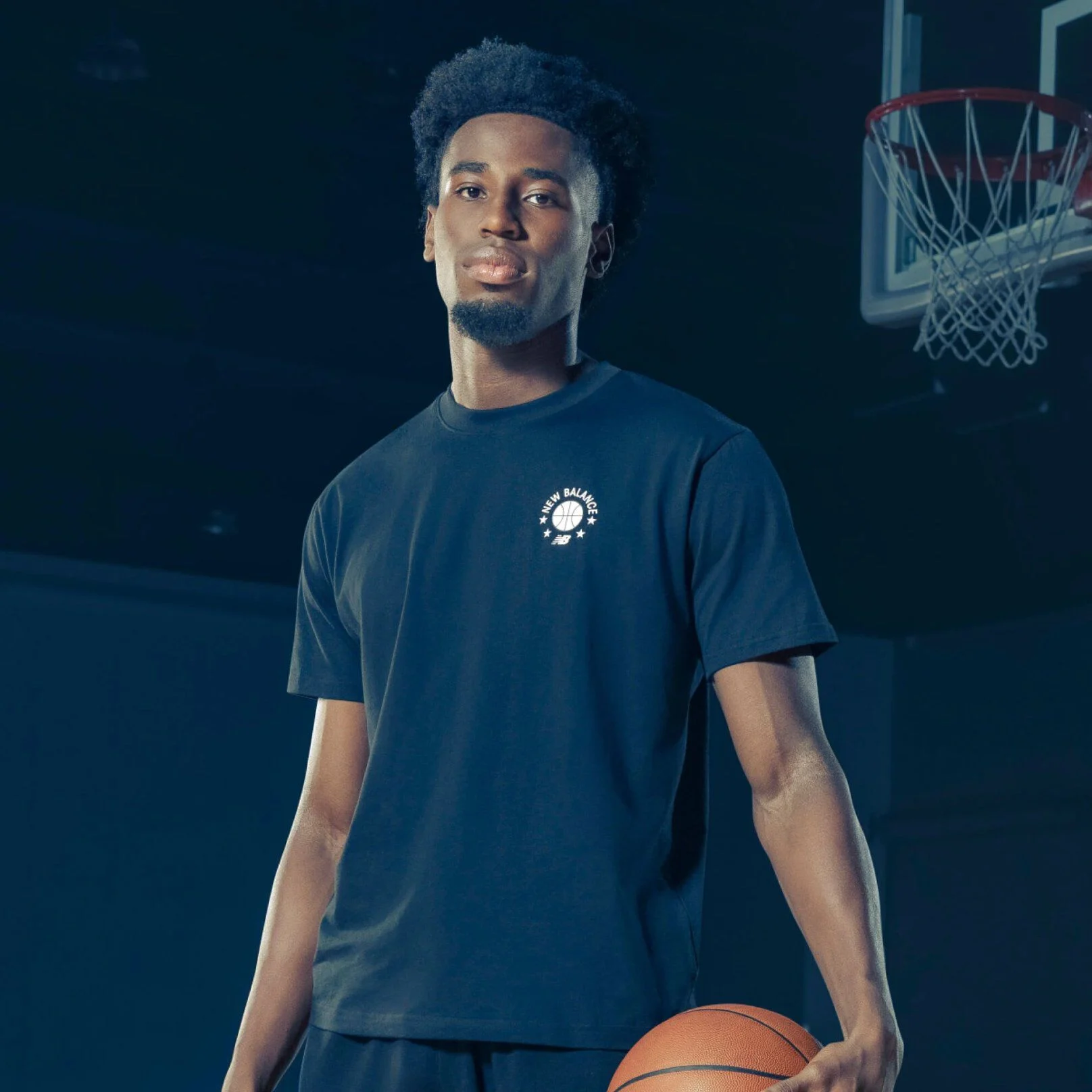A person stands on an indoor basketball court wearing a dark T‑shirt with a small circular New Balance Basketball graphic on the chest, holding a basketball at their side.