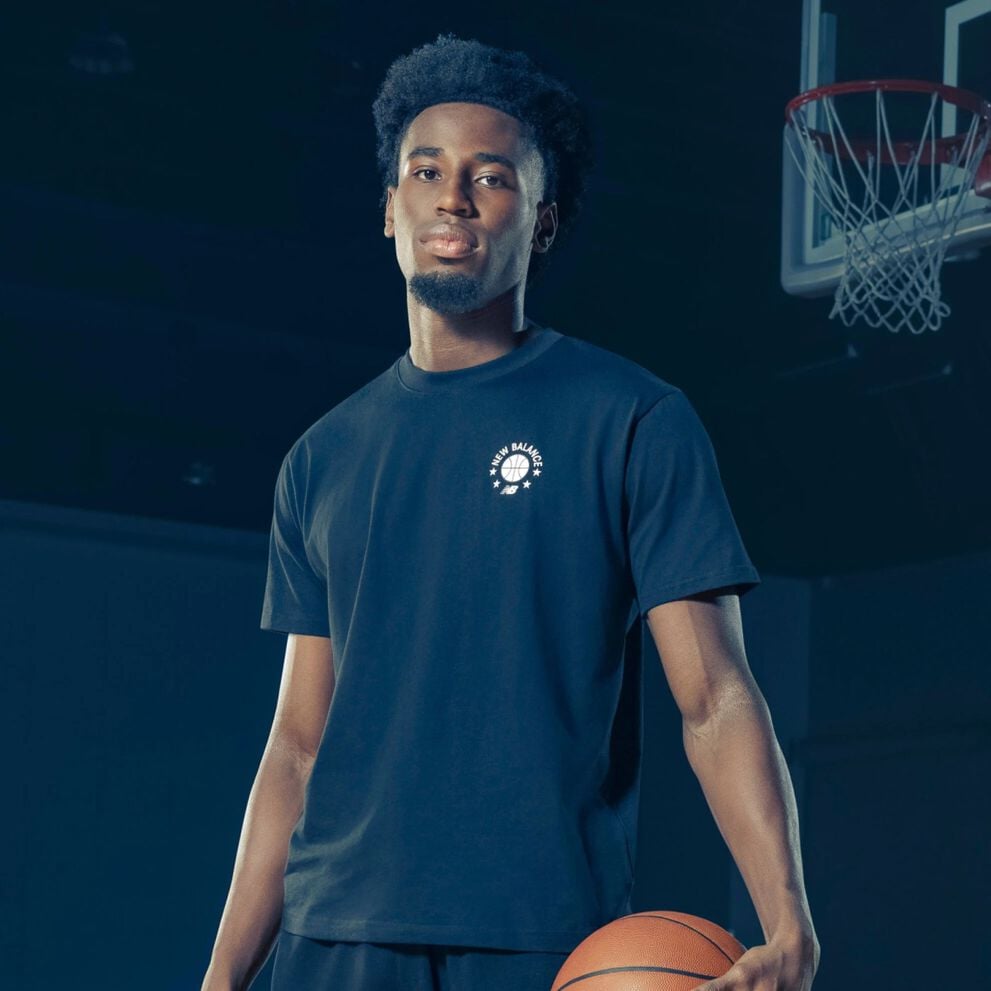A person stands on an indoor basketball court wearing a dark T‑shirt with a small circular New Balance Basketball graphic on the chest, holding a basketball at their side.