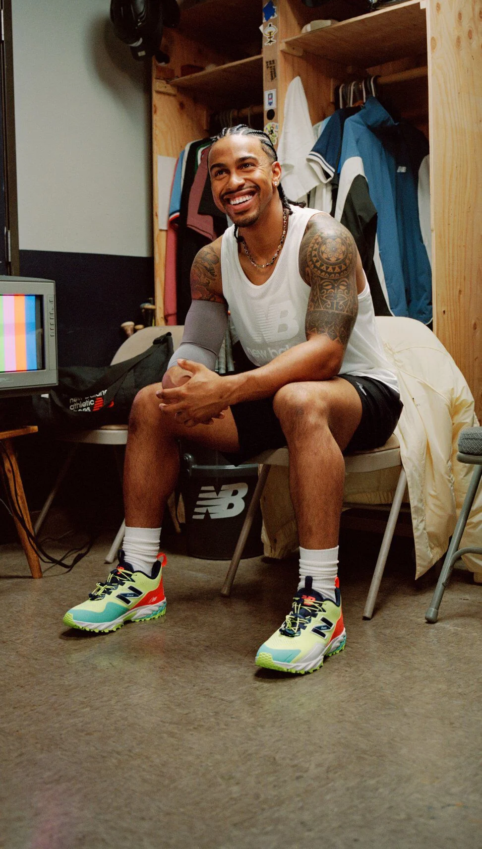 A person sitting in a locker room wearing brightly colored New Balance athletic shoes with neon green, orange, and black detailing, paired with a sleeveless top, shorts, and white socks.