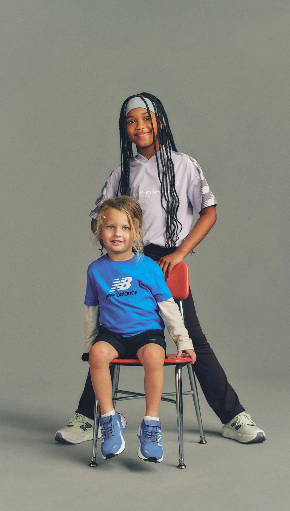 Two children in New Balance outfits pose with one seated on a chair in blue sneakers and the other standing behind with a hand on the chair against a neutral gray background.