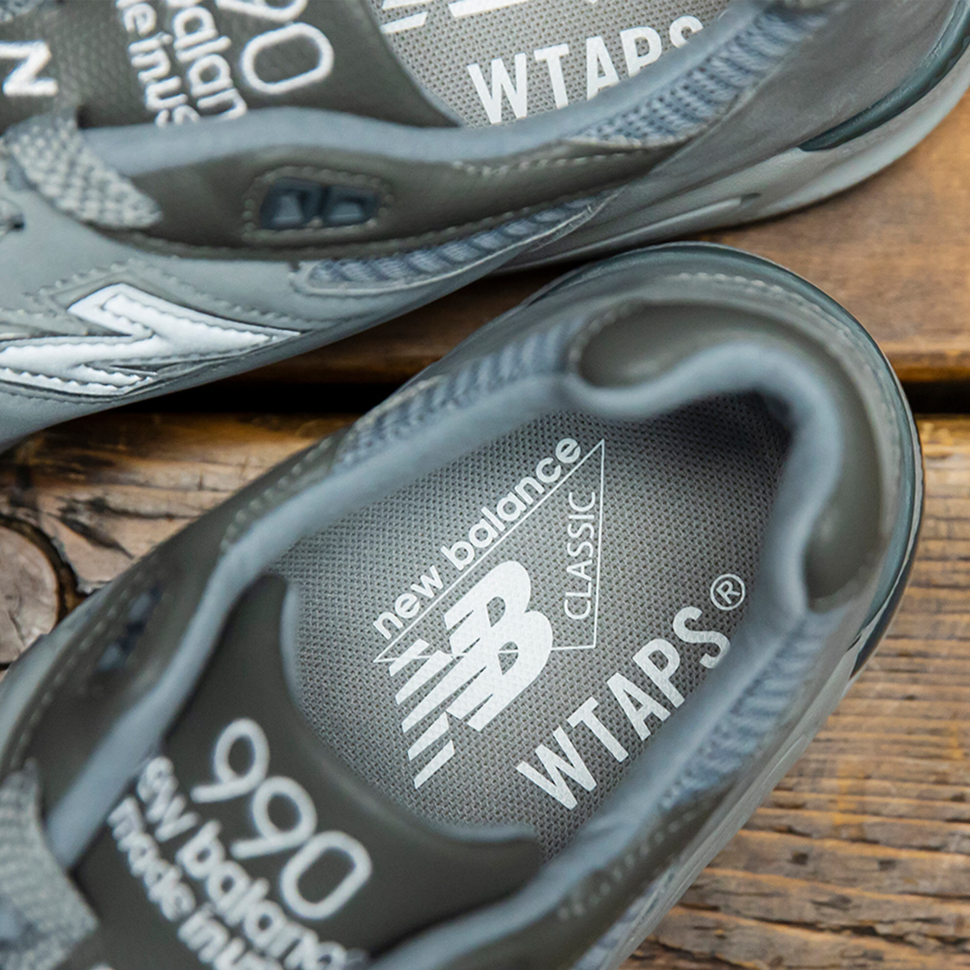 Close-up overhead image focused on the insole branding of a grey NB x WTAPS 990v2 sneaker sitting on rustic wood flooring