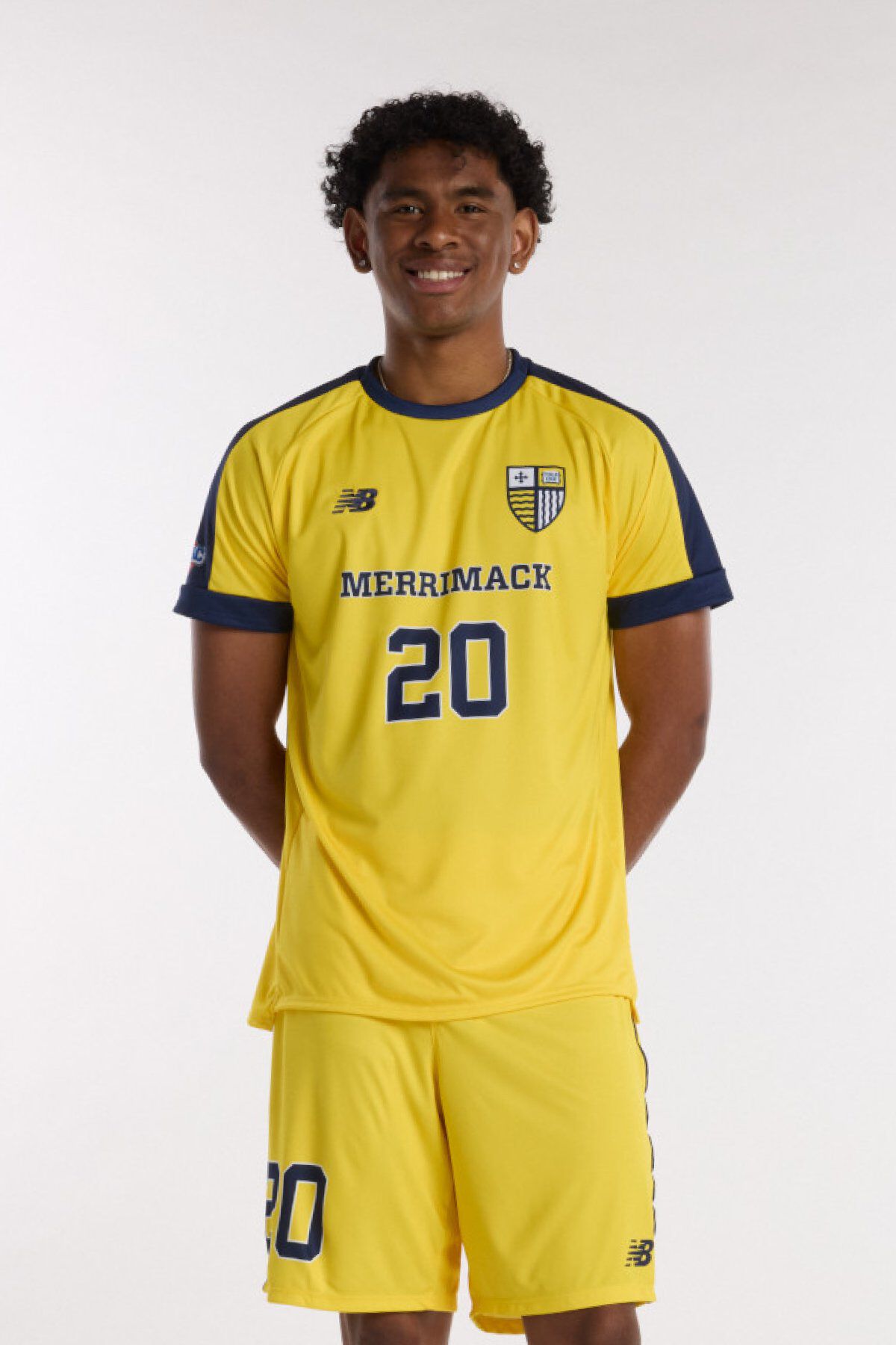 Young man wearing a yellow Merrimack soccer jersey with number 20 and matching shorts, standing against a light background.