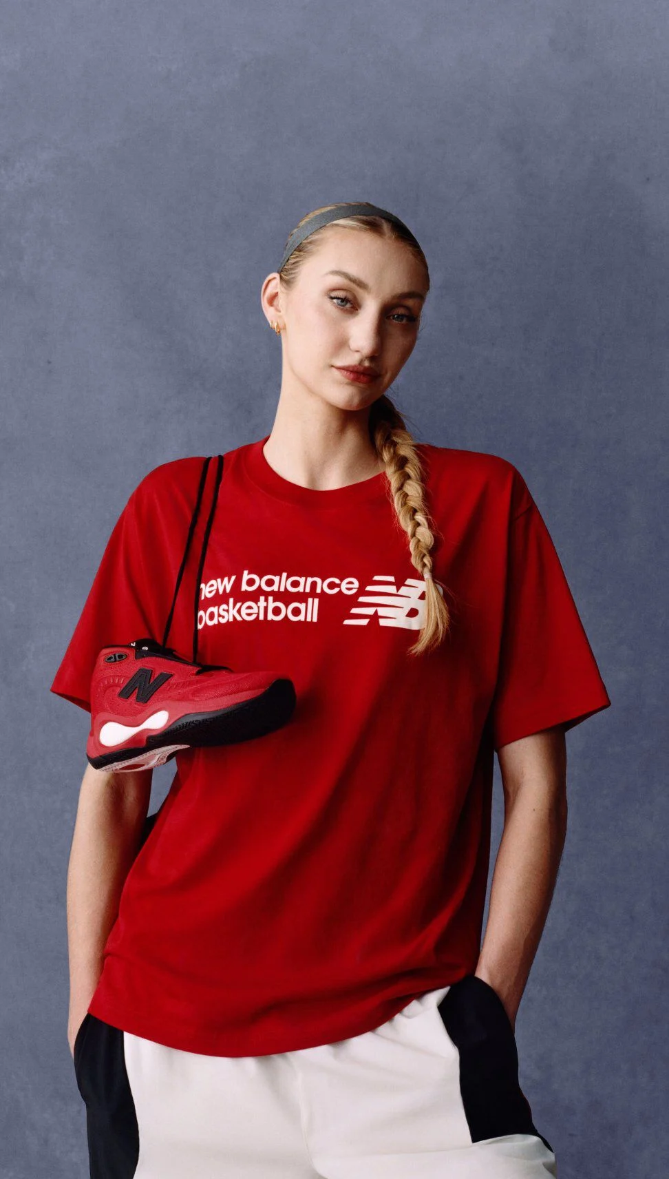 Athlete wearing a red New Balance basketball T-shirt holding a matching red and black sneaker against a gray studio background, with the &lsquo;new balance basketball&rsquo; logo visible on the shirt.