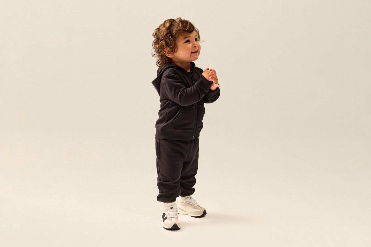 Toddler wearing a black hooded sweatsuit and white New Balance sneakers standing in a clean studio setting.