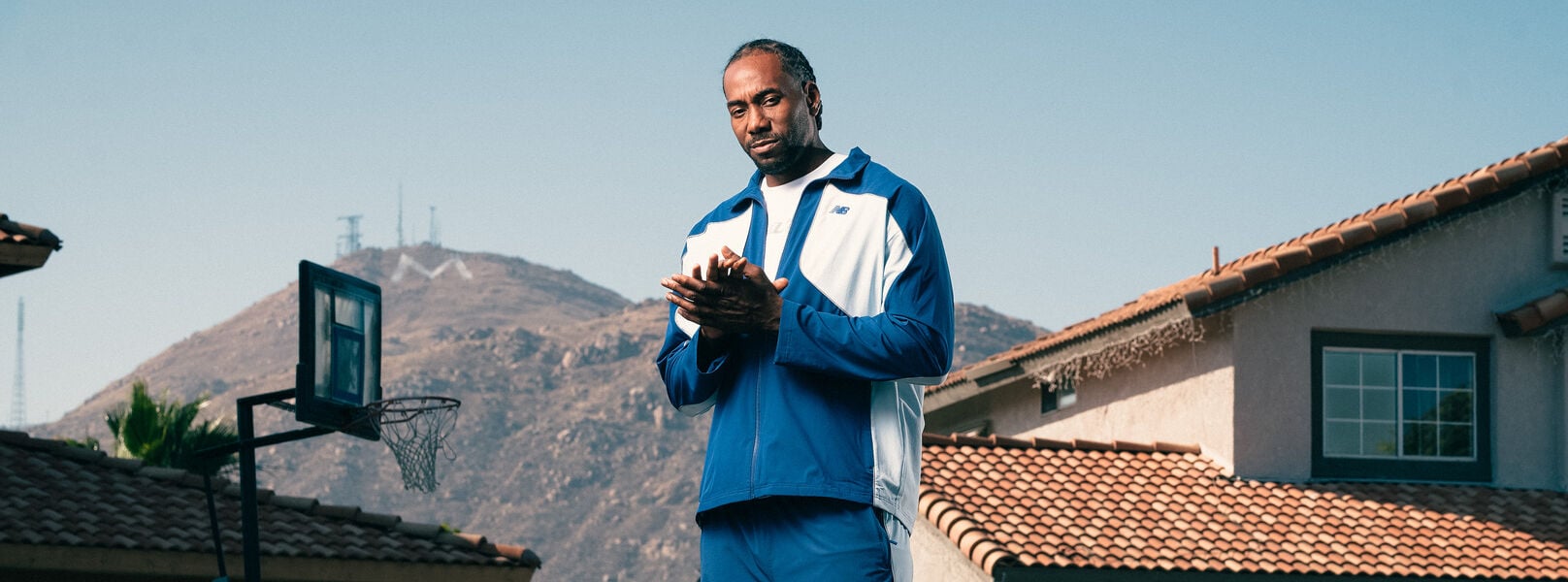 Person wearing a blue and white tracksuit and matching sneakers stands on a suburban street with mountains and houses in the background.