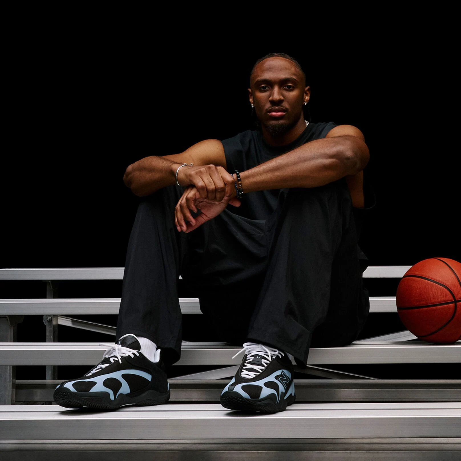 Person seated on metal bleachers beside a basketball, highlighting black-and-light-blue athletic sneakers in a dark studio setting.