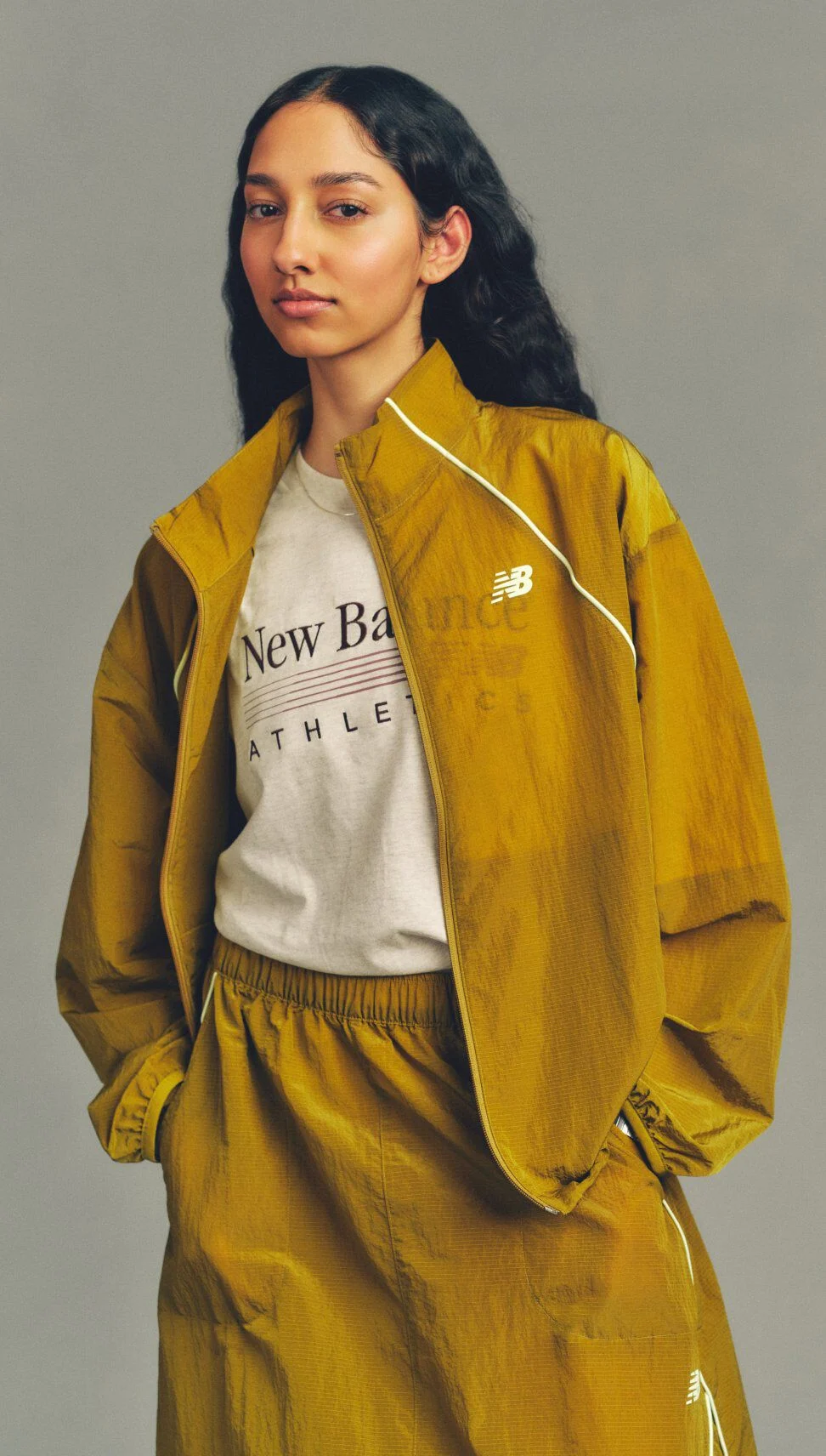 Close-up studio image of a person wearing a mustard-yellow New Balance windbreaker and matching pants over a graphic T-shirt against a neutral gray background.