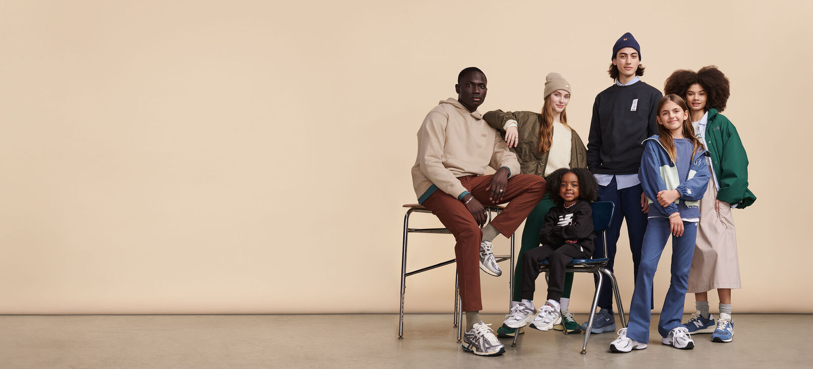 Group of men, women and kids wearing New Balance clothing and shoes posed around school desk against beige background in studio environment 