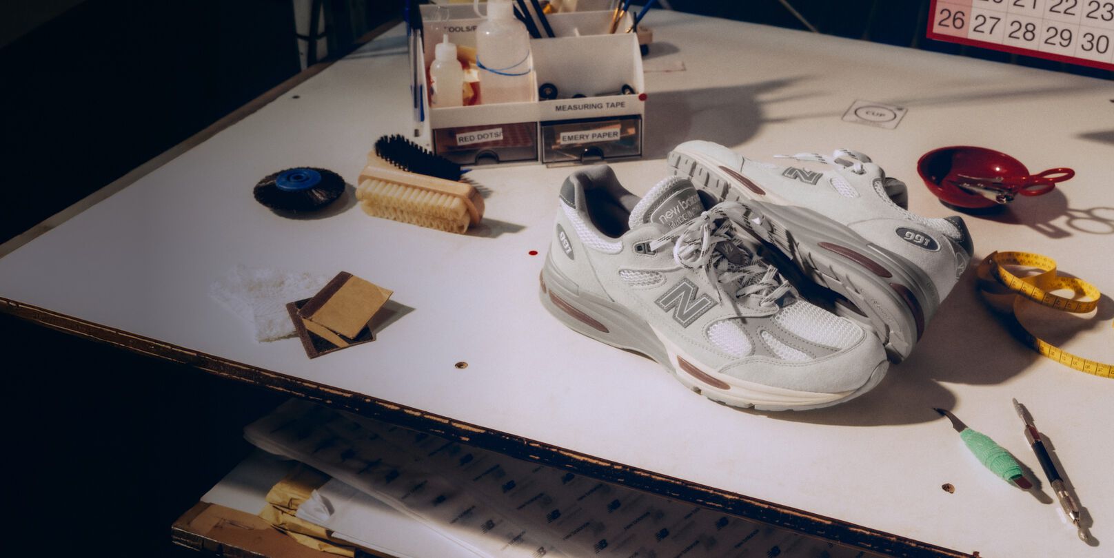 Un par de tenis Made in UK de New Balance sobre un banco de trabajo blanco, rodeado de herramientas para la fabricaci&oacute;n de calzado y otros equipos textiles.