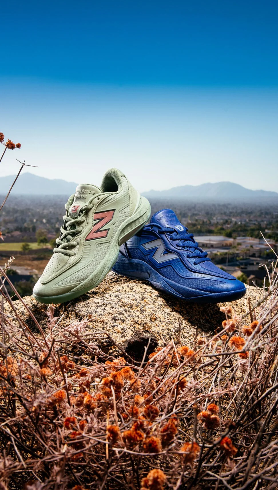 Dos zapatillas New Balance, de color verde y azul, en una ladera rocosa con vistas a un paisaje de ciudad y monta&ntilde;as.