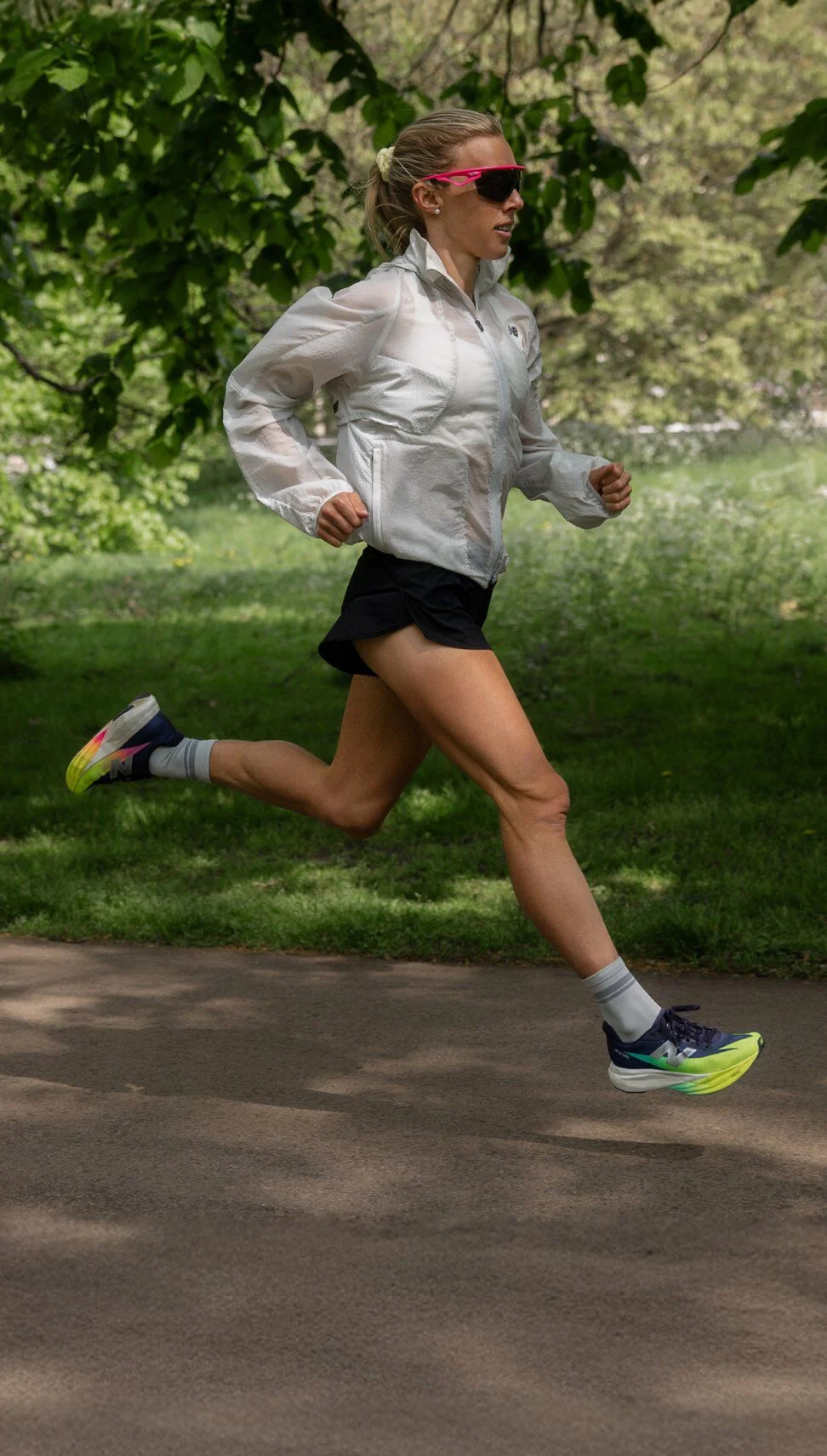 Emily Sisson running outdoors on a paved path in a park, wearing a white jacket, black shorts, and brightly colored running shoes. 