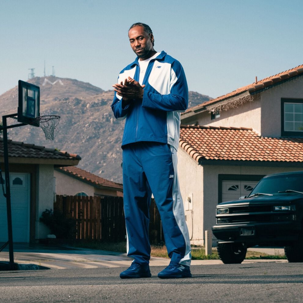 Person wearing a blue and white tracksuit and matching sneakers stands on a suburban street with mountains and houses in the background.