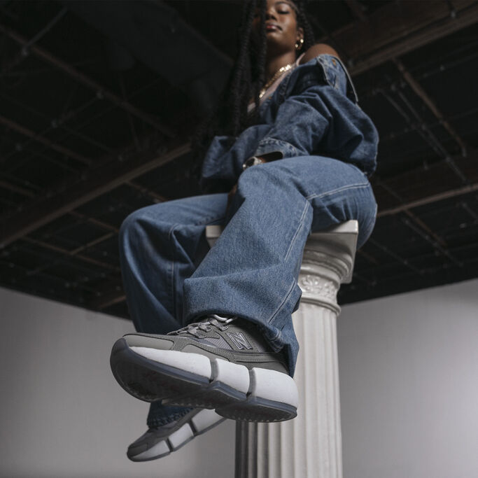 Women in a denim jacket, jeans, and grey Vision Racers sitting on a stone pedestal with foot kicked out towards camera.
