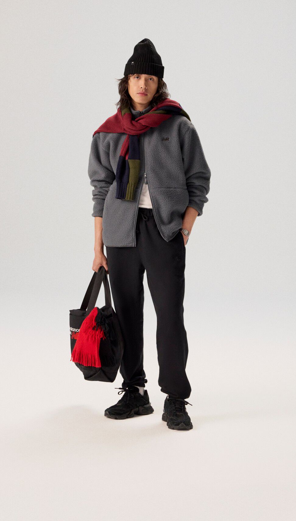 Joven de pie sobre un fondo gris con una chaqueta gris sosteniendo una bolsa.