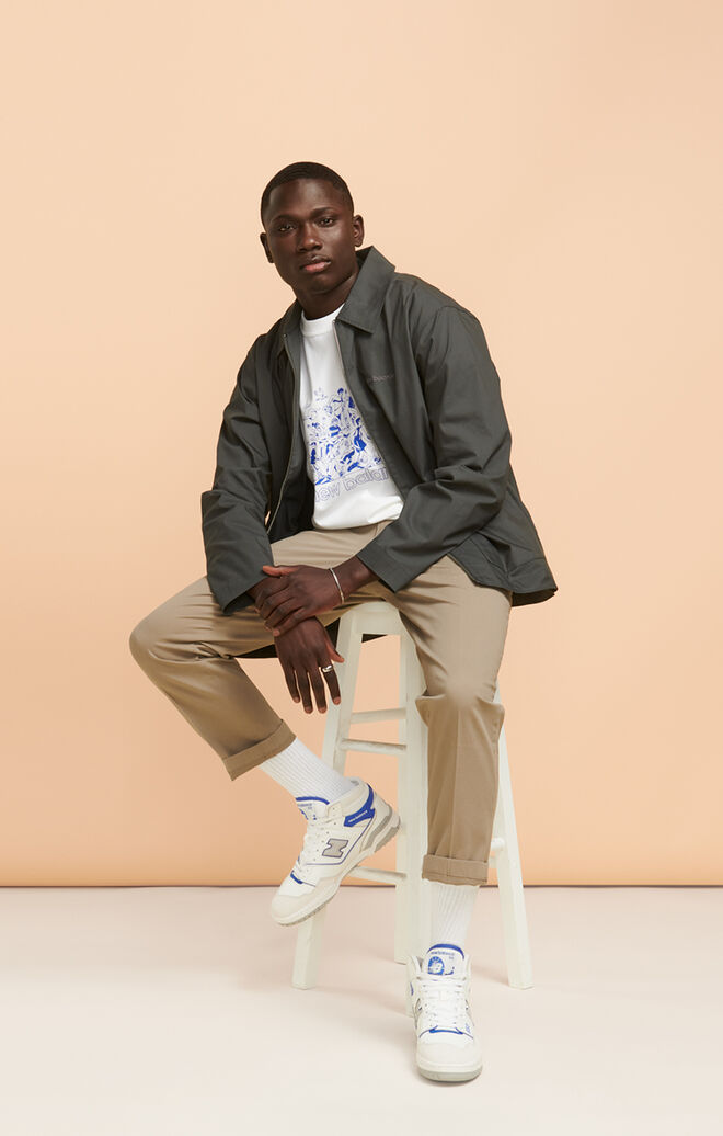 A young man posing on a stool wearing a NB graphic tee with khaki pants and white NB 650 sneakers with blue accents