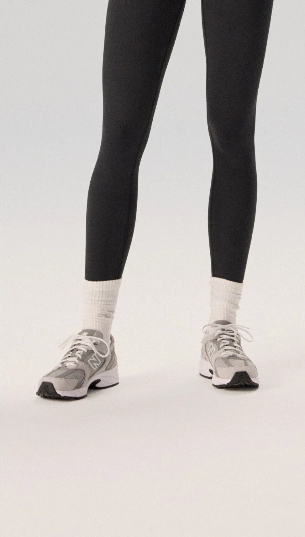 Mujer con medias negras y zapatillas de correr grises sobre un fondo gris.