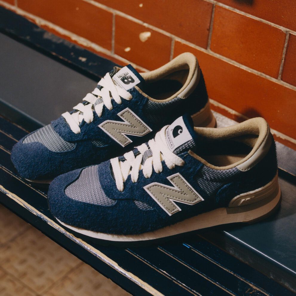 Pair of blue Carhartt New Balance 990v1 shoes on a bench in a boxing gym.