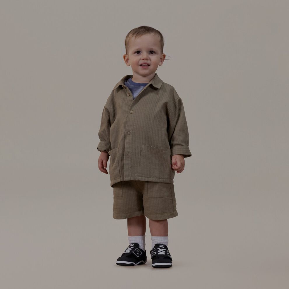 Bebé con un traje color canela de pie sobre un fondo gris.