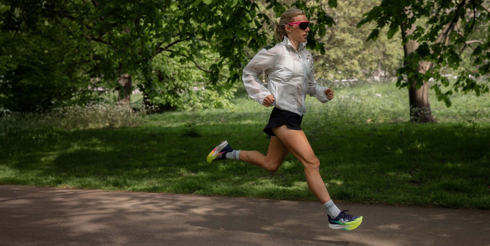 Emily Sisson corriendo al aire libre en un camino pavimentado en un parque usando una chaqueta blanca, pantalones cortos negros y calzado de correr con colores brillantes.&nbsp;