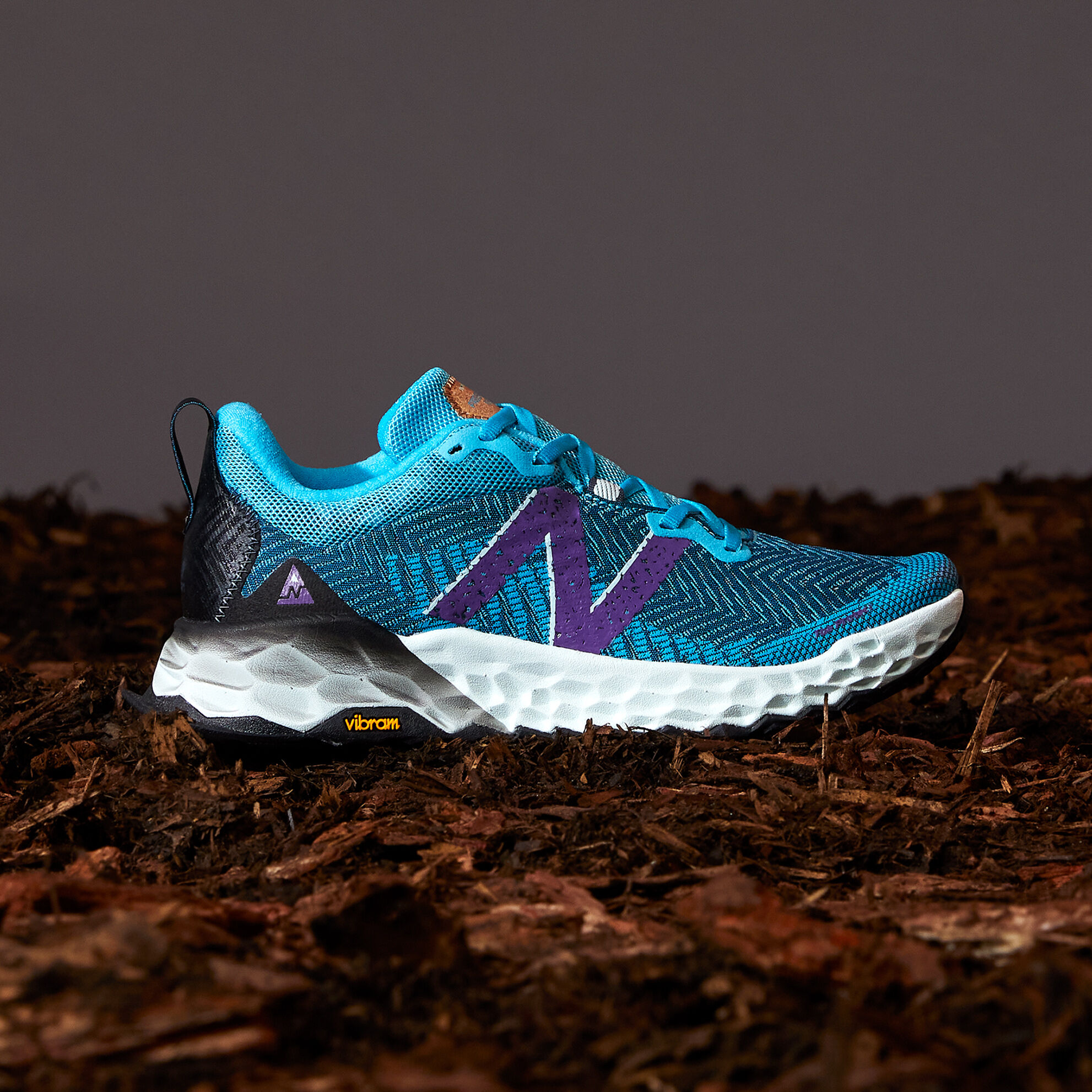 Bright blue women’s trail shoe on dirt and sticks with a dark grey background. 
