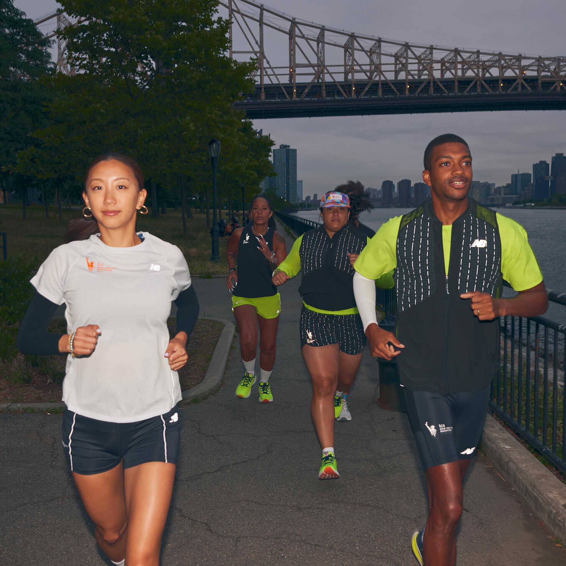 A group of individuals run towards the camera wearing New Balance clothing with reflective accents and the TCS New York City Marathon logo.