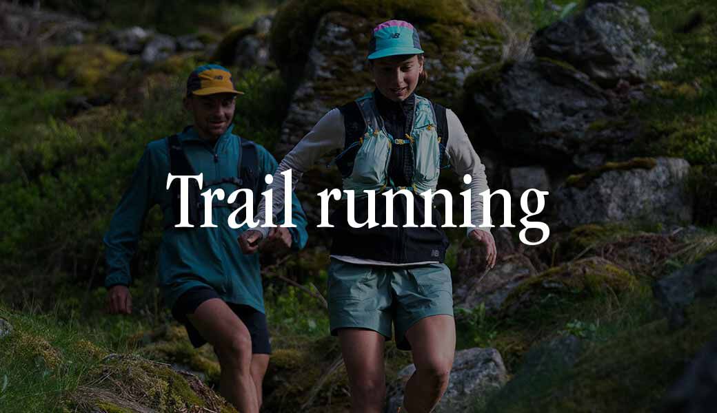Two runners in New Balance running apparel wearing trail running shoes for men and navigating a lush green forest trail, surrounded by dense trees and foliage.