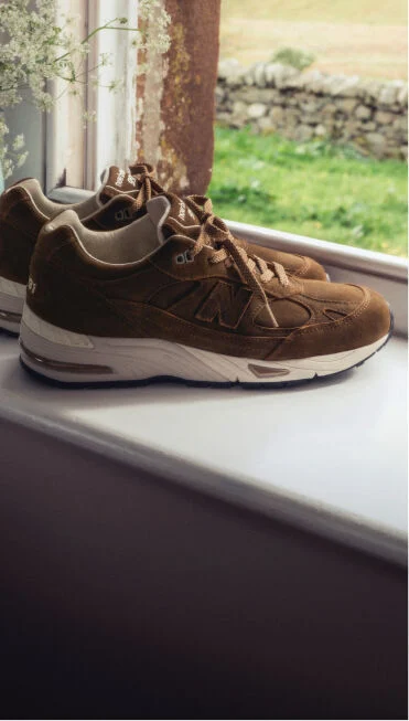 A pair of New Balance Made in UK sneakers on a windowsill next to a small vase of white flowers.