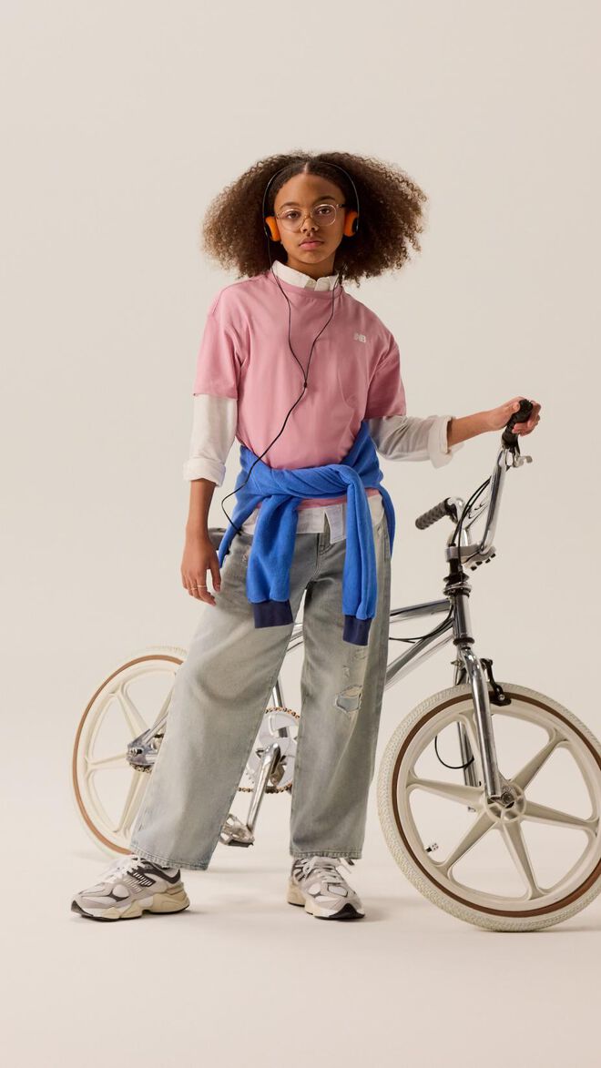 Young kid standing in front of a bike wearing blue jeans and a pink shirt. 