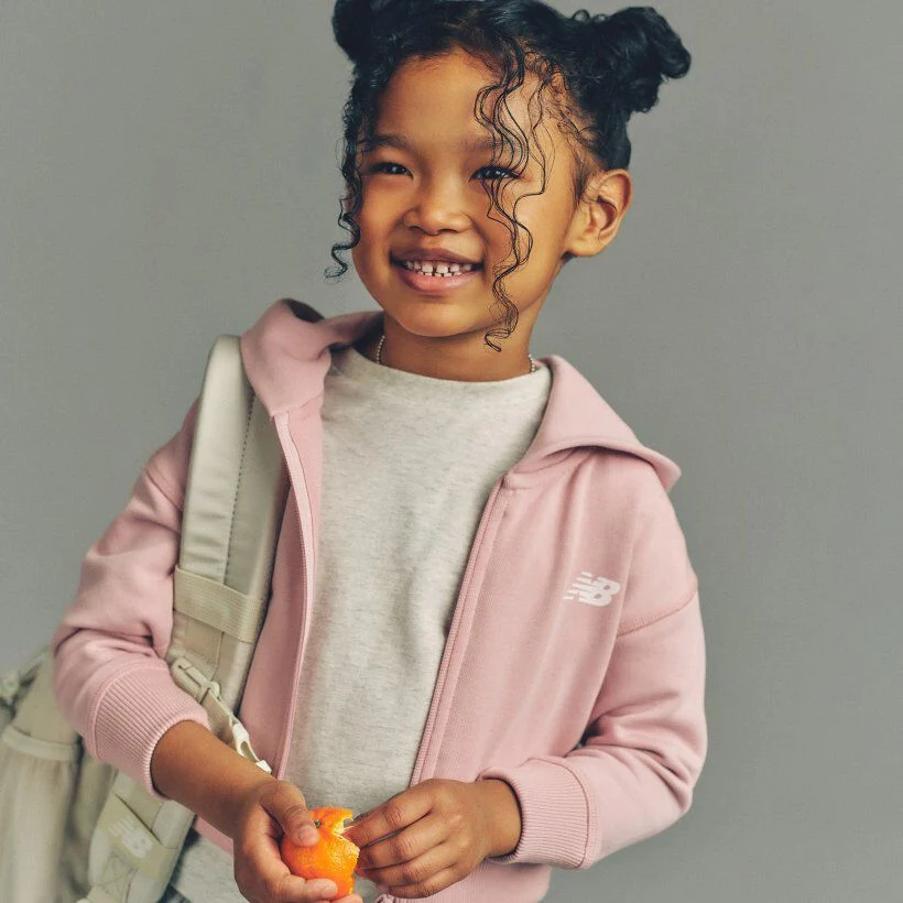 Child wearing a pink New Balance hoodie and carrying a backpack, holding a small orange while standing against a plain background.