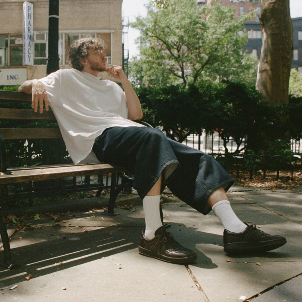 Jack Harlow sitting outside on a park bench wearing dark brown New Balance sneakers.