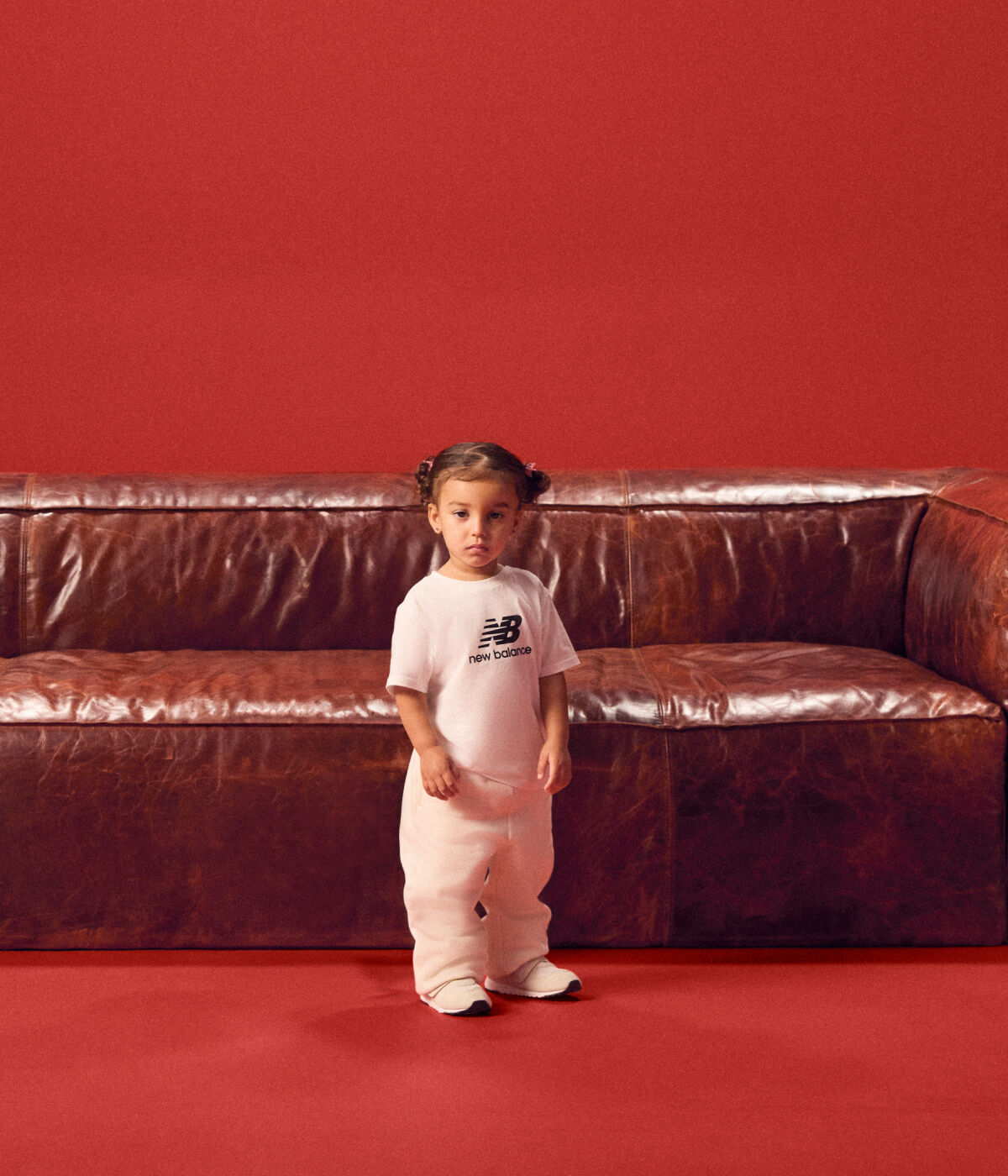 Baby standing in front of a red couch wearing a white tshirt.