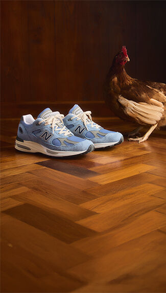 Un par de zapatillas New Balance Made in UK colocadas sobre un suelo rústico de madera en espiga con un pollo de pie junto a ellas.  