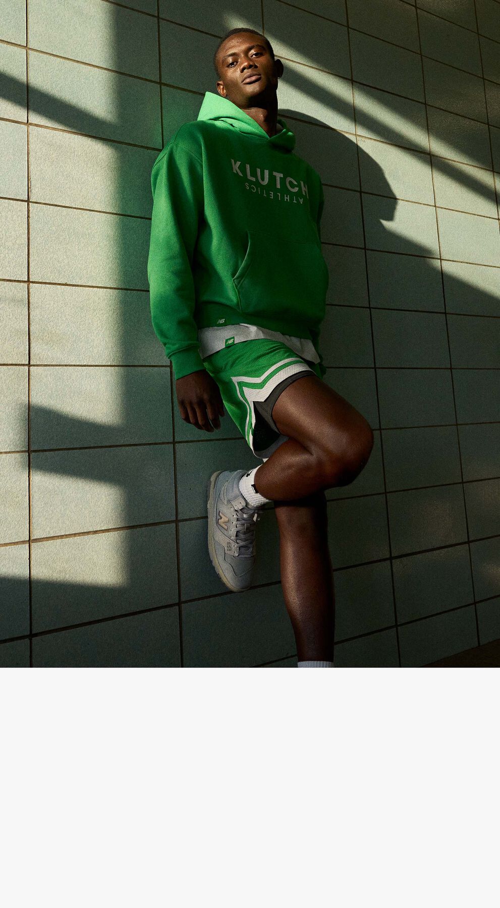 Man wearing Klutch Athletics by New Balance, green hoodie and green shorts posing against a wall with dramatic lighting 