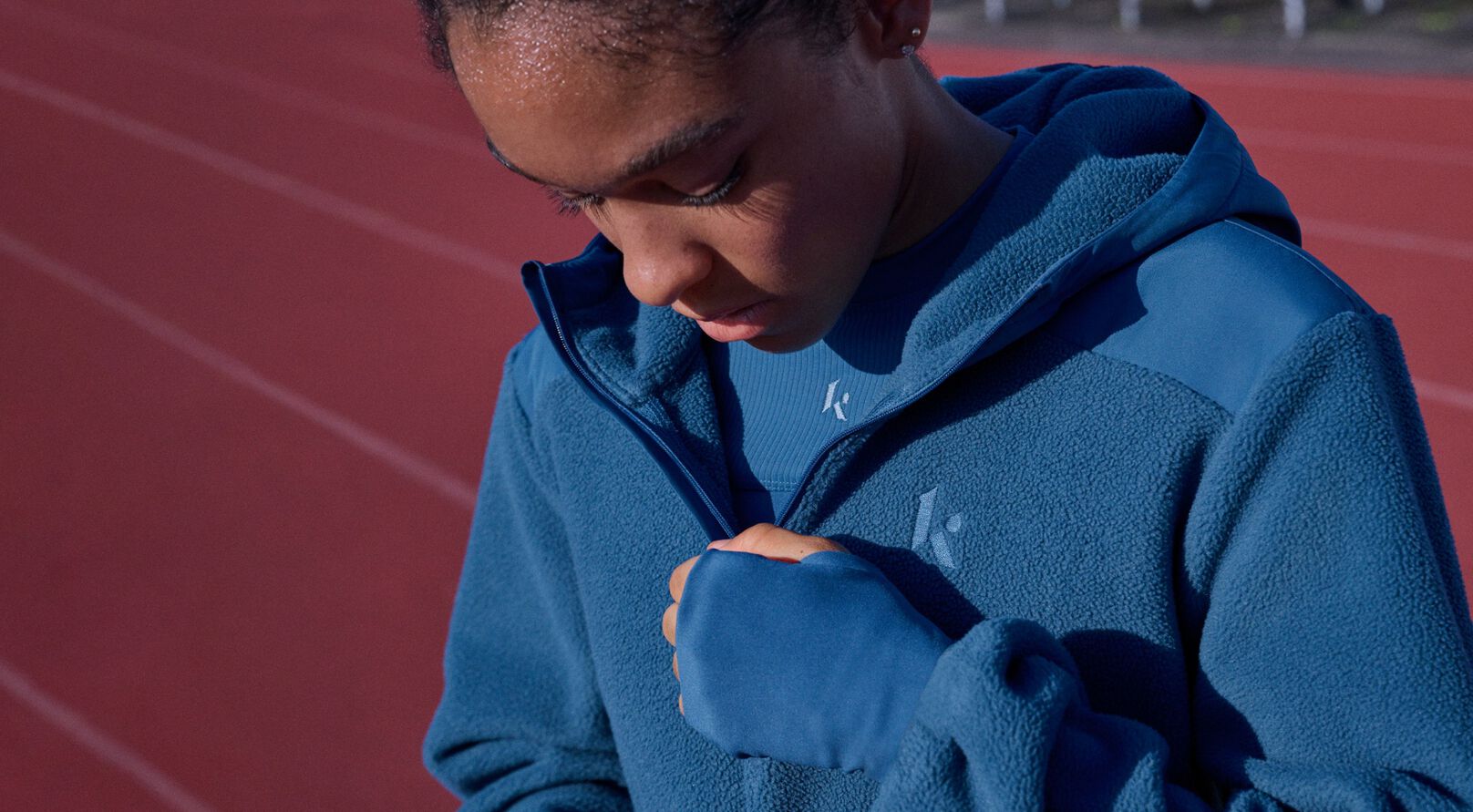 Close-up of an athlete wearing a blue fleece jacket and performance top on a running track.